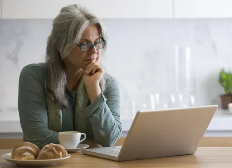 A woman at her kitchen counter, thinking over what is on her laptop