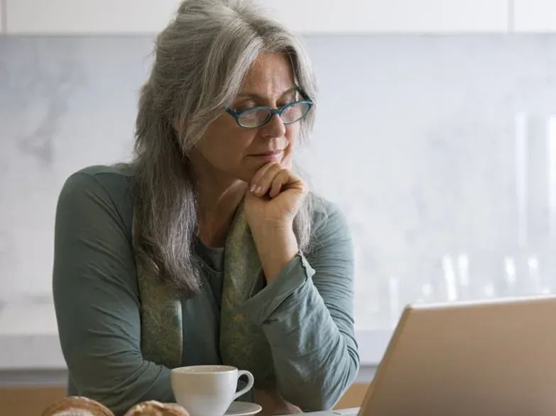 A retired woman at her kitchen table, thinking over what is on her laptop screen